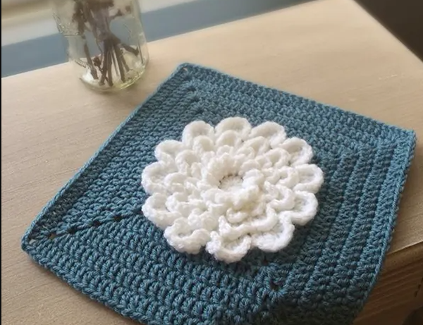 A blue crochet square with white flower on a table with glass vase. 