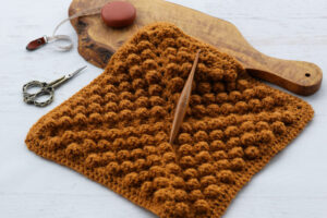 a crochet quilt block in honey yellow yarn with a crochet hook, scissors and measuring tape