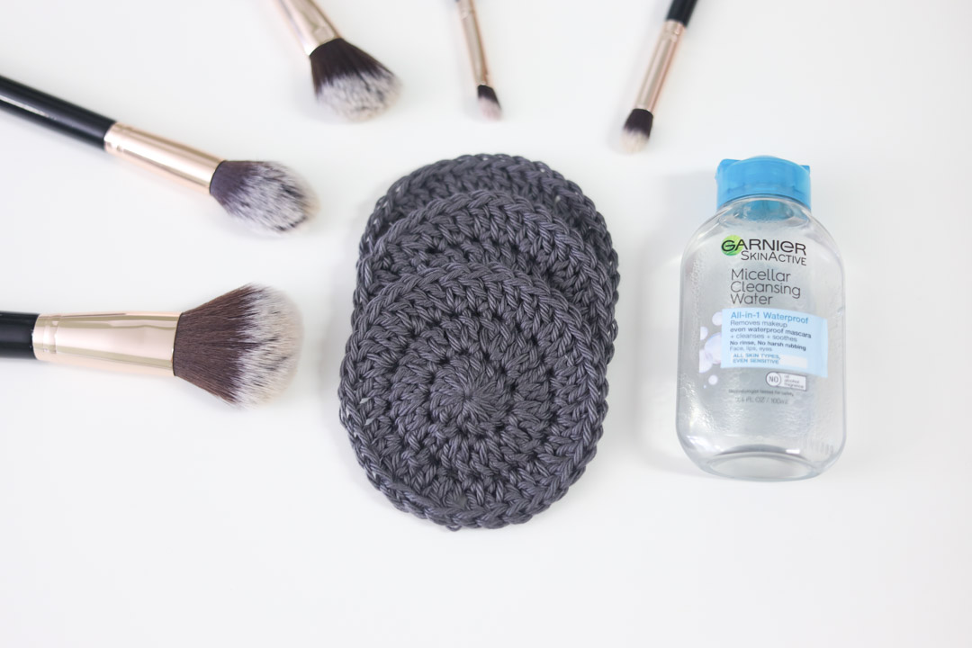 a stack of gray crochet face scrubbies with makeup brushes and micellar water