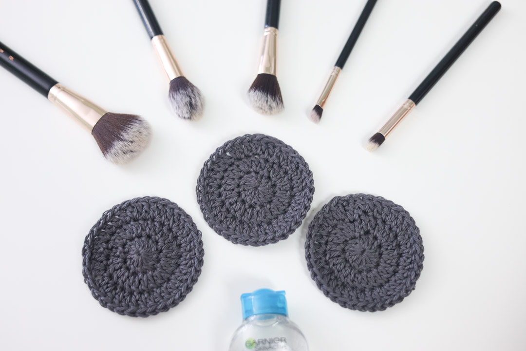 3 circular crochet face scrubby rounds on a white surface with makeup brushes and a bottle of micellar water