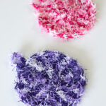 two crochet dish scrubbies made with scrubby yarn in purple and pink on a white surface