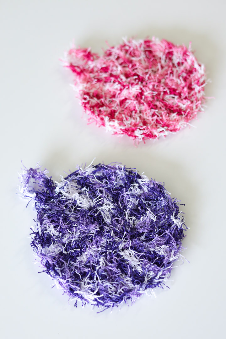 two crochet dish scrubbies made with scrubby yarn in purple and pink on a white surface