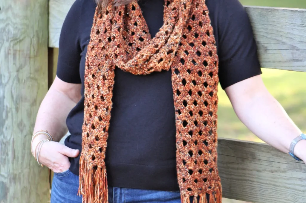 Woman standing next to a wooden fence wearing a crochet variegated burnt orange scarf.