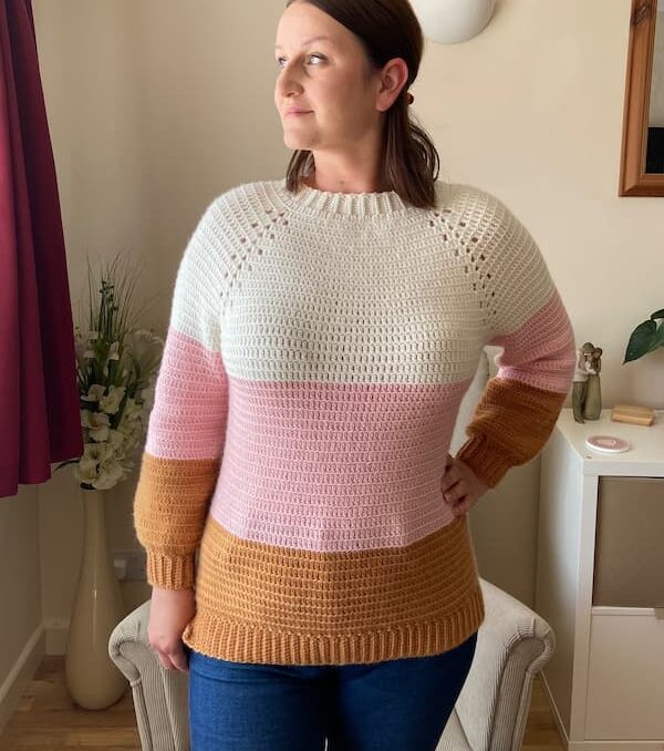 A woman standing in front a chair wearing a crochet color block sweater in cream, light pink and golden tan.