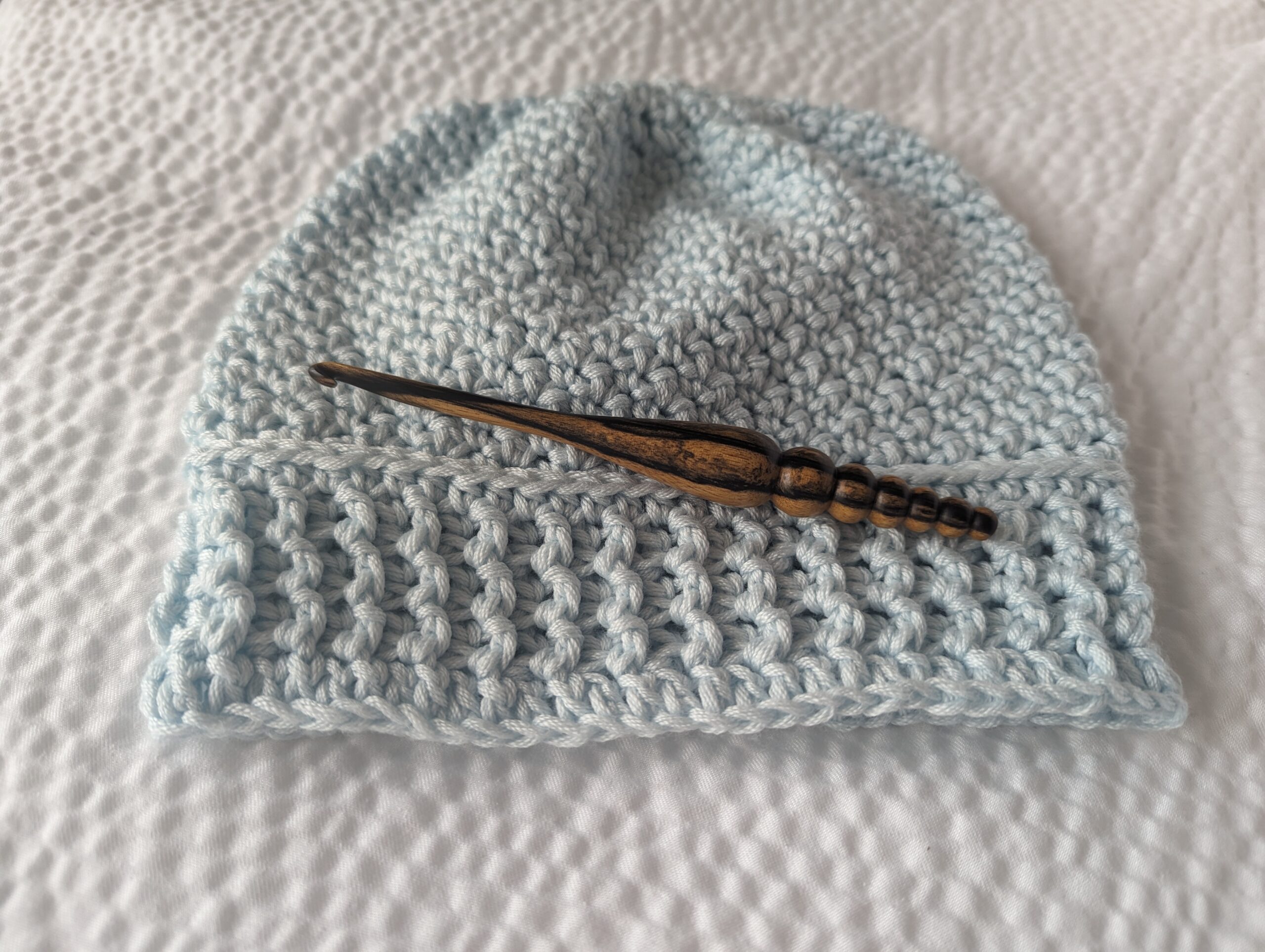 a blue crochet beanie with a wood crochet hook on a white blanket
