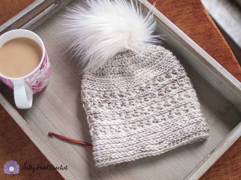 highly textured cream crochet beanie with fur pom on a gray serving tray with a cup of coffee and a crochet hook placed on a wooden table.