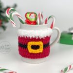 A white coffee mug with a santa themed mug cozy on it and with candy canes inside it