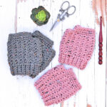 A set of gray crocheted fingerless gloves and pink crocheted fingerless gloves placed next to a wooden crochet hook, scissors and a small plant on a white wooden table.